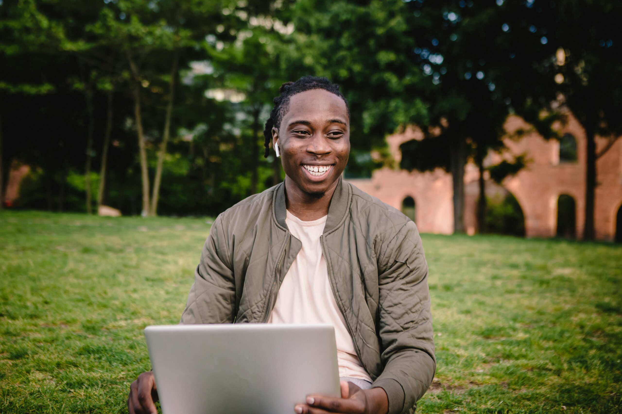 Positive African American male in casual clothes with wireless earbuds using laptop and smiling while sitting on green lawn and enjoying free time in quiet park